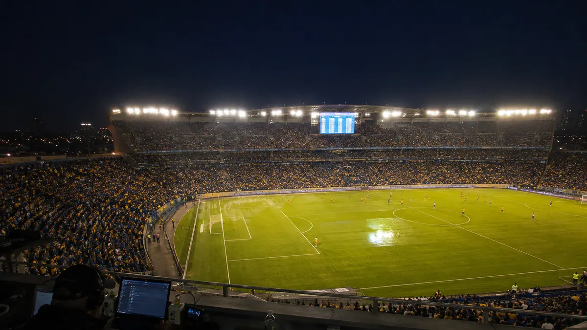 A press-box view over a floodlit league pitch at night with an electronic table board glowing in the far stand