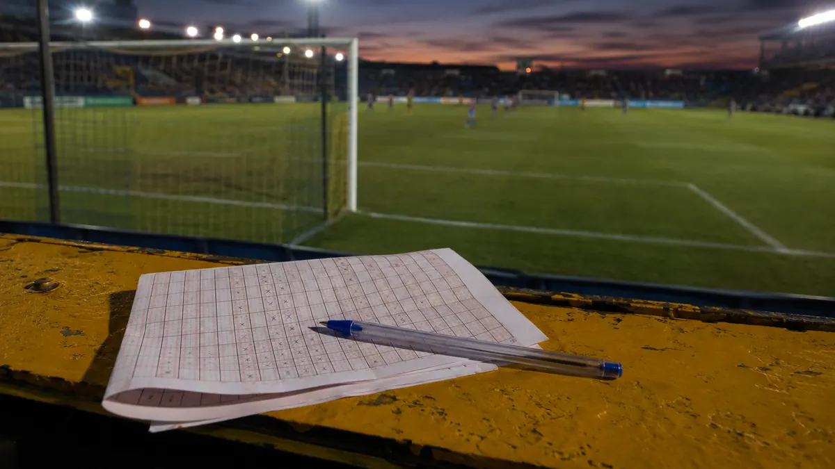 A paper pools coupon and a pen on the front-row barrier of a floodlit South African stadium at dusk